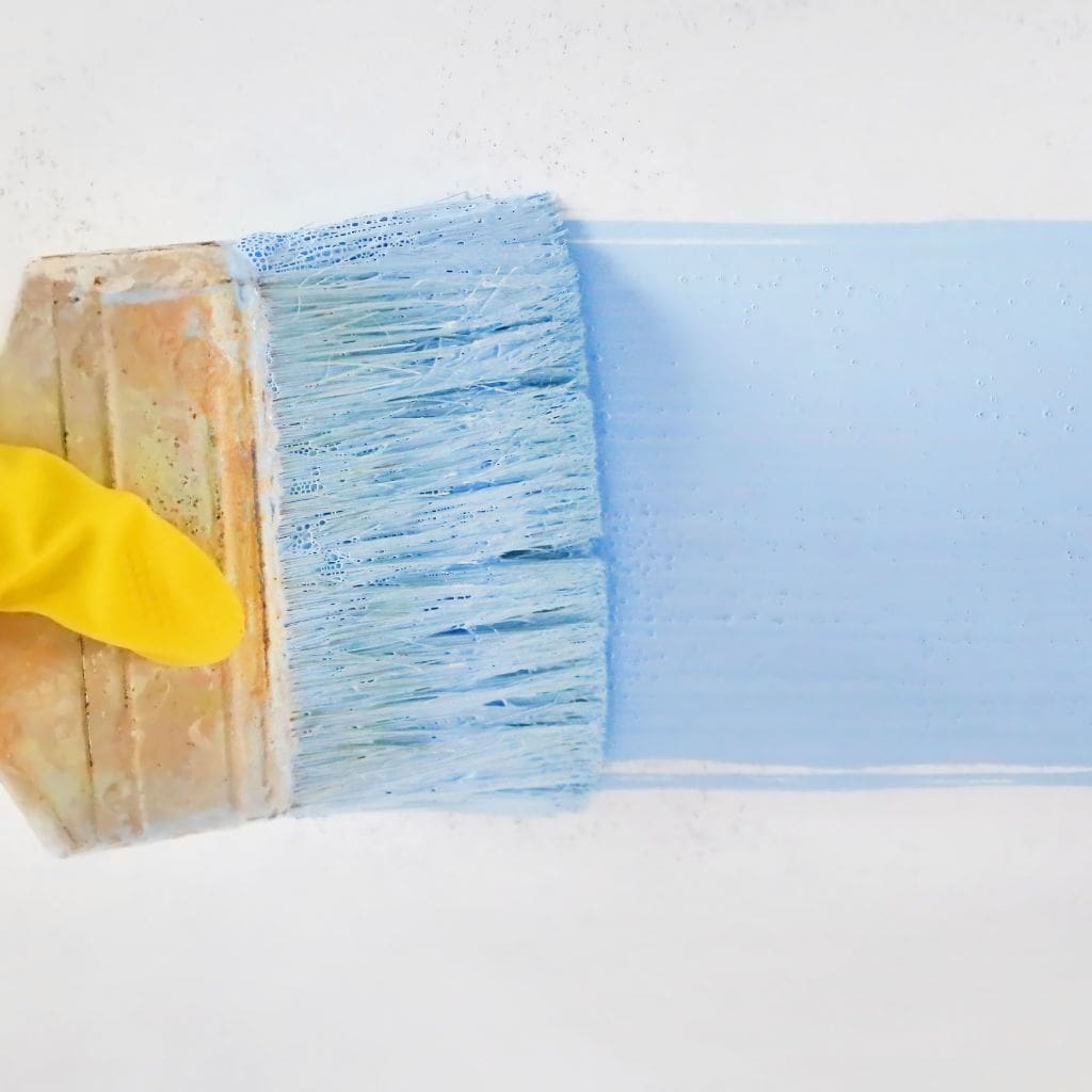 Person holding a paint brush painting a stroke of light blue paint on a white wall.