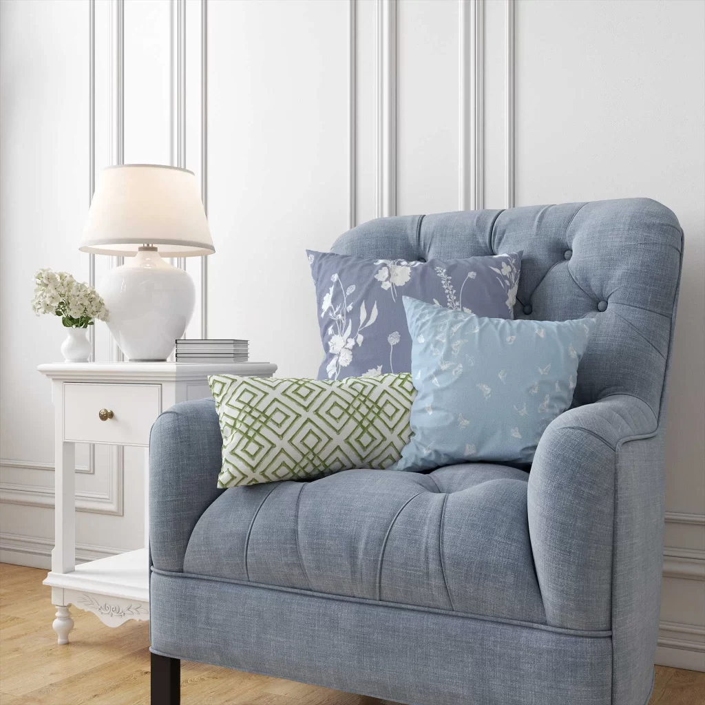 A blue linen armchair with three Color Caravan throw pillows in a white paneled room with a white side table and white table lamp.